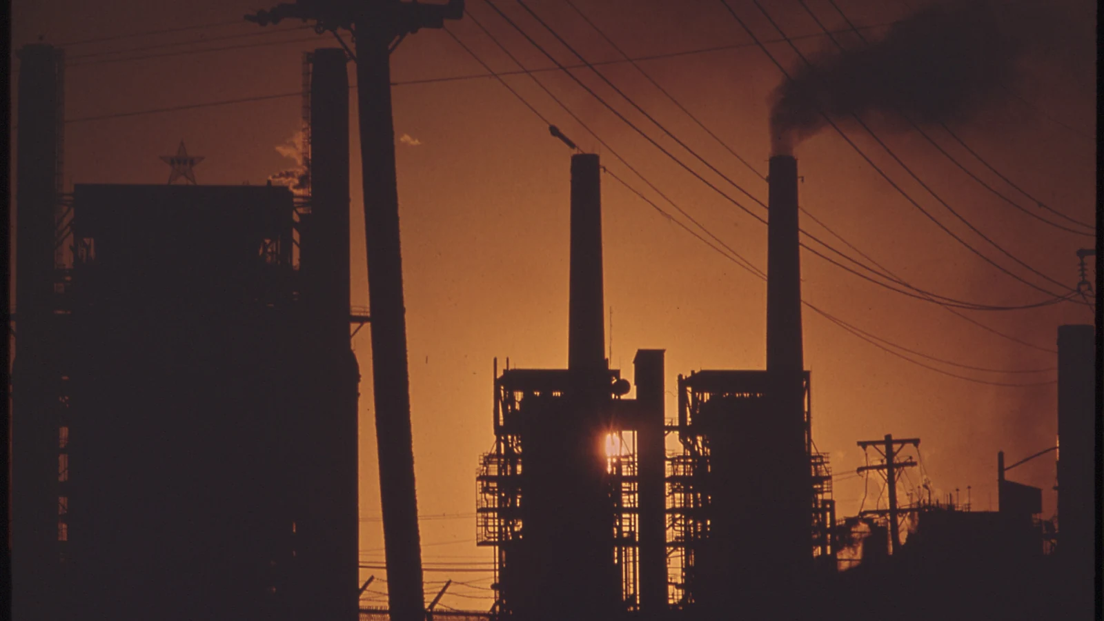 An oil refinery appears in silhouette against an orange sunset.
