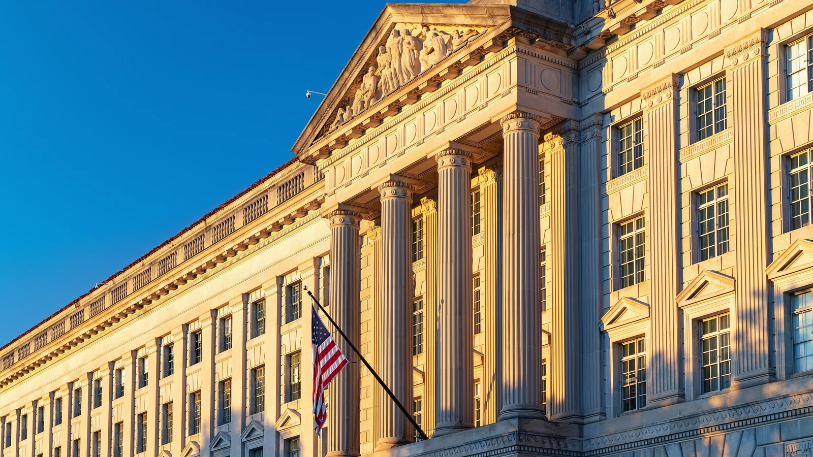 The Herbert C. Hoover Building in Washington glows in late-day sunlight beneath a blue sky.
