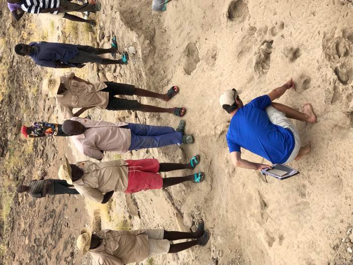 Researcher Kevin Hatala crouches on an excavated track surface and points toward shallow hominin footprints.