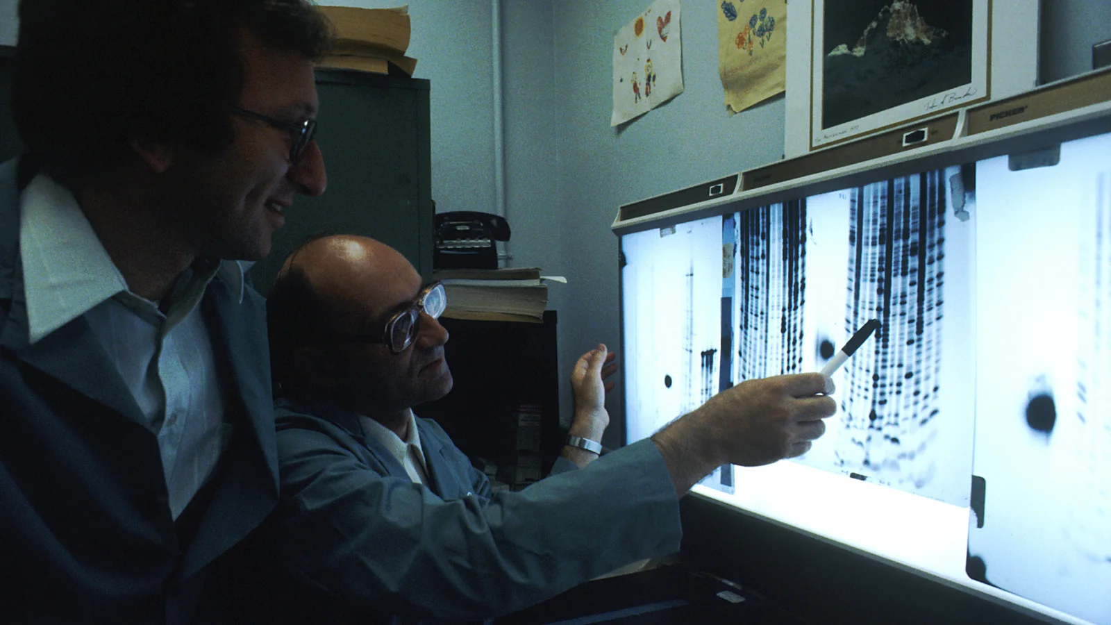 Two scientists in lab coats examine illuminated DNA sequence information in a laboratory.