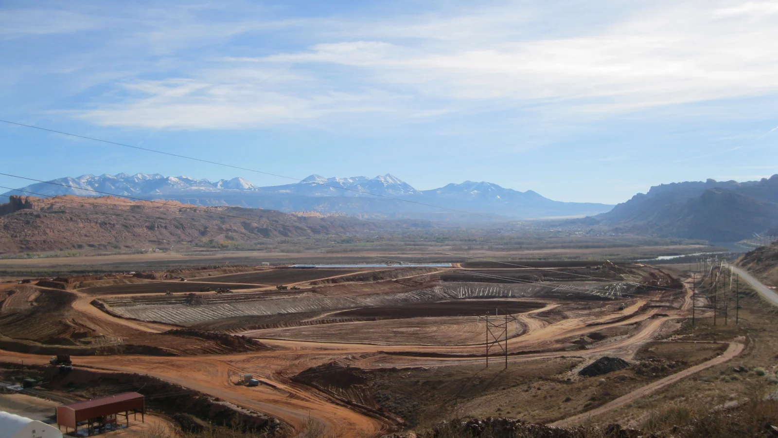 A broad uranium-tailings remediation site with excavation roads below red cliffs, snowy mountains and the Colorado River corridor near Moab.