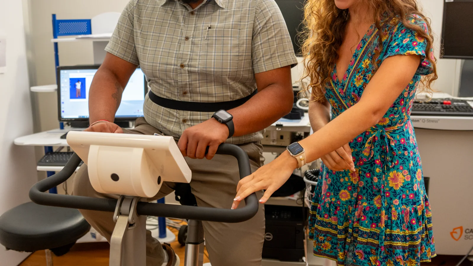 A person pedals a stationary exercise bike while wearing monitoring equipment in a bright clinical research room.