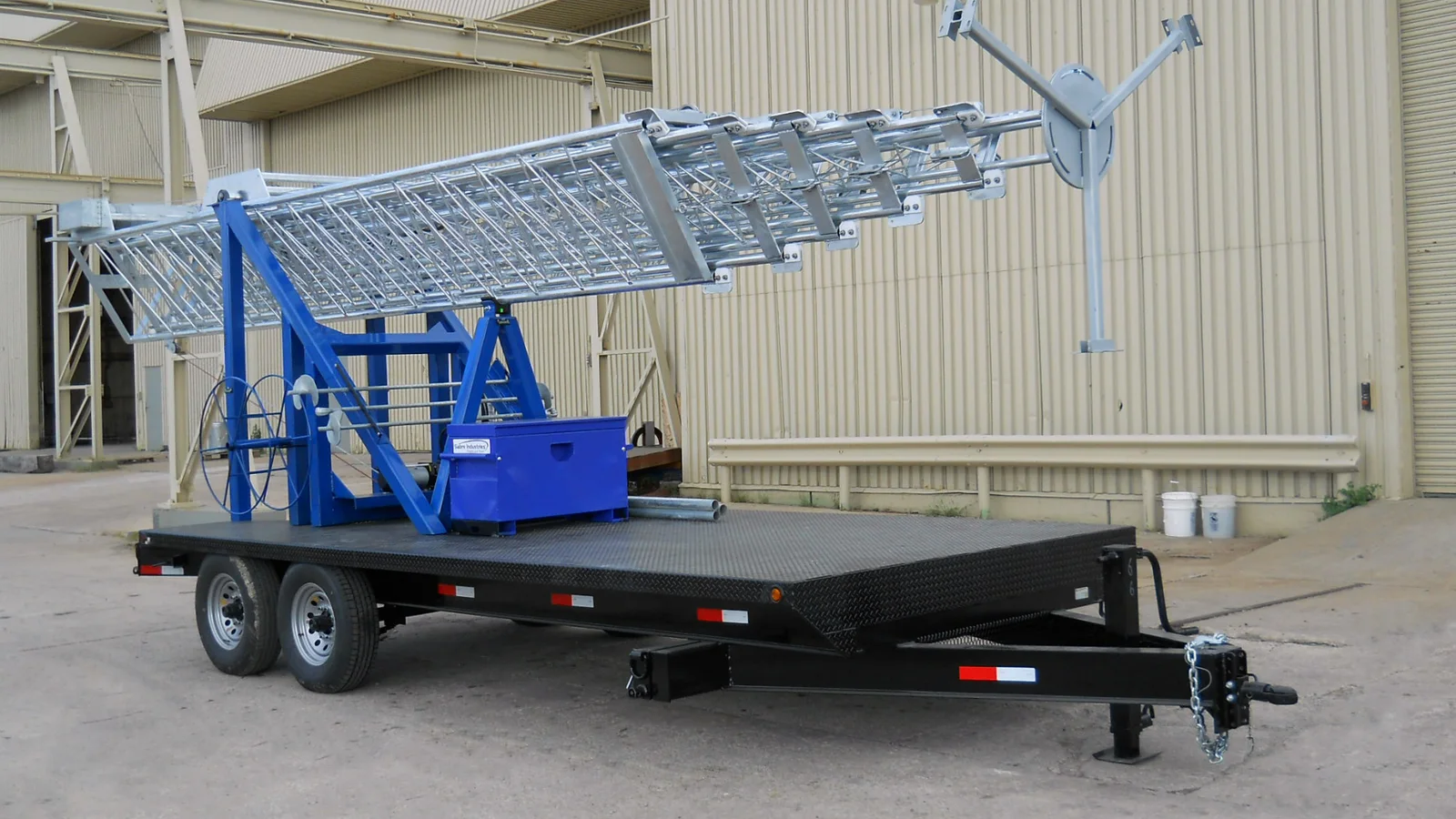 A folded lattice mobile telecommunications tower rests on a dual-axle black trailer outside an industrial building.