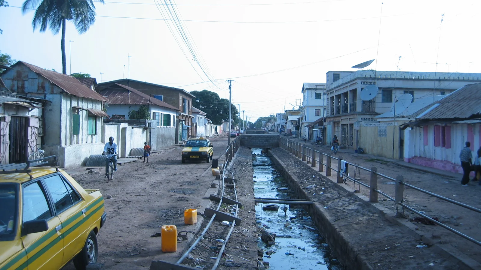 Cars and pedestrians move along a broad downtown street in Banjul lined with low commercial buildings and trees.