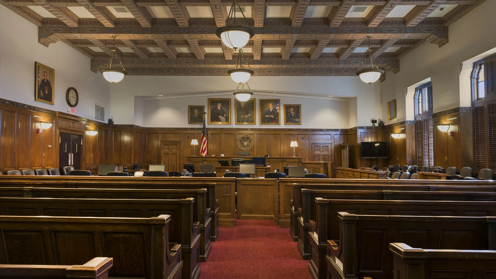 Wood benches face the judge's bench in a formal federal courtroom with tall windows and pendant lights.