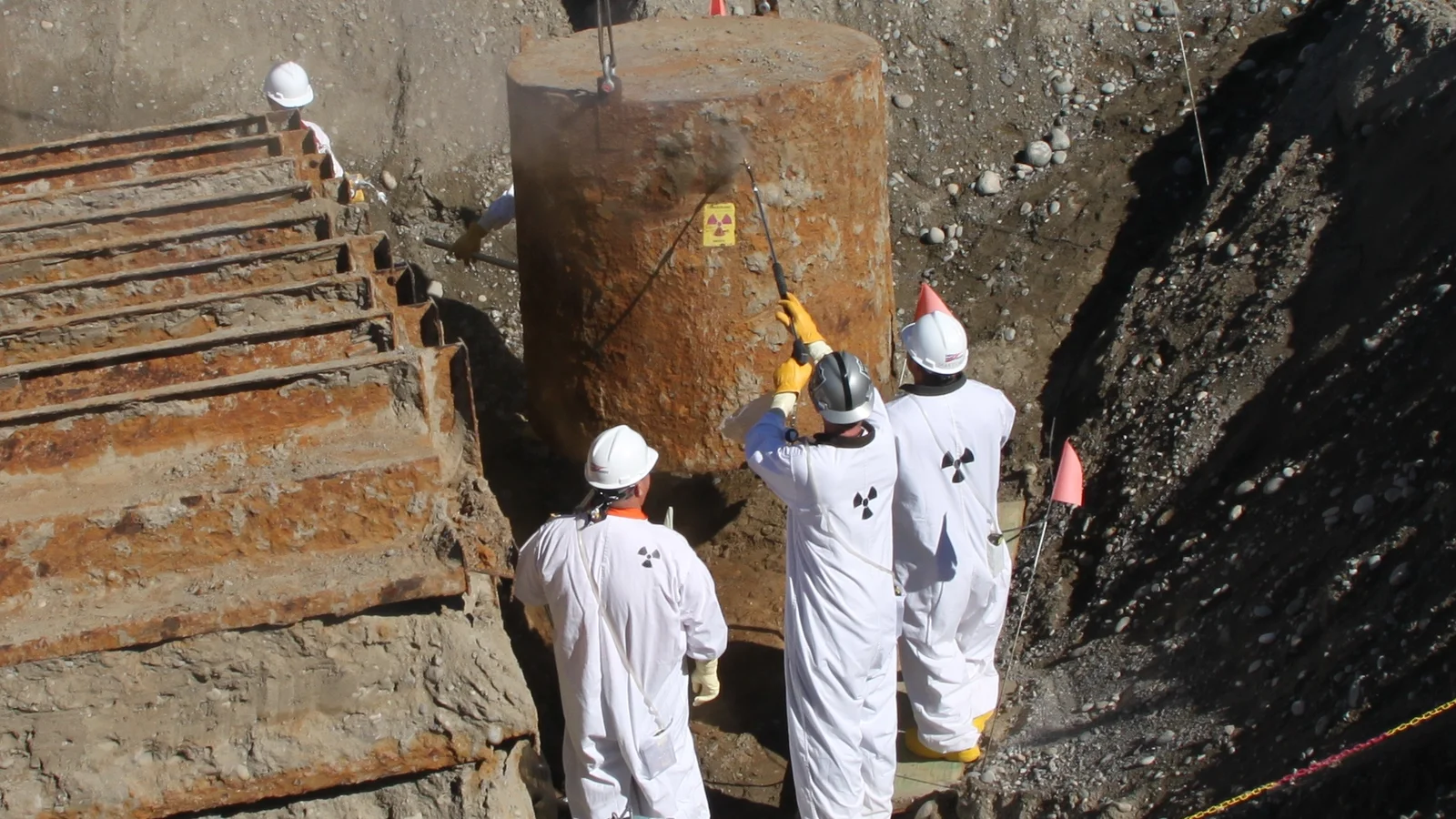 Workers in protective gear operate heavy equipment at a fenced nuclear cleanup site.