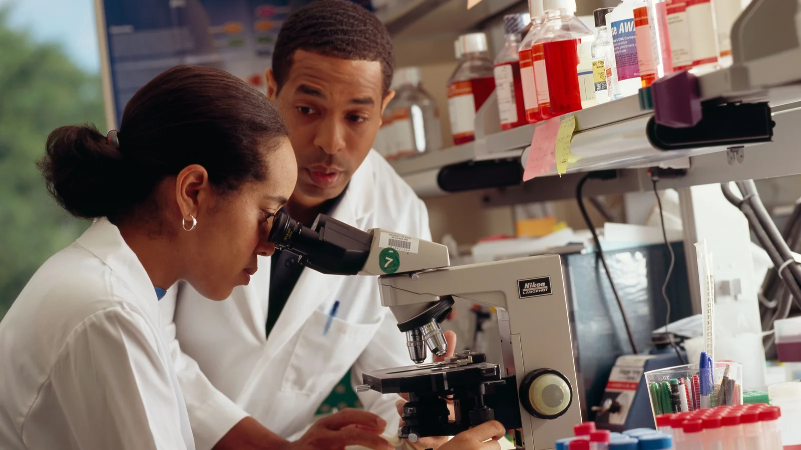 One researcher looks through a microscope while a second researcher observes beside her in a laboratory.