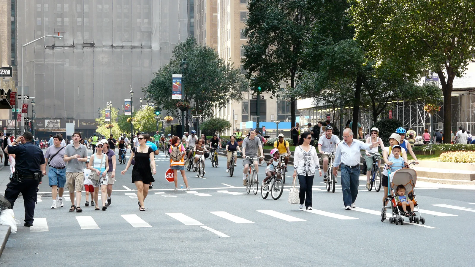 A large crowd of pedestrians and cyclists fills a broad New York City avenue.