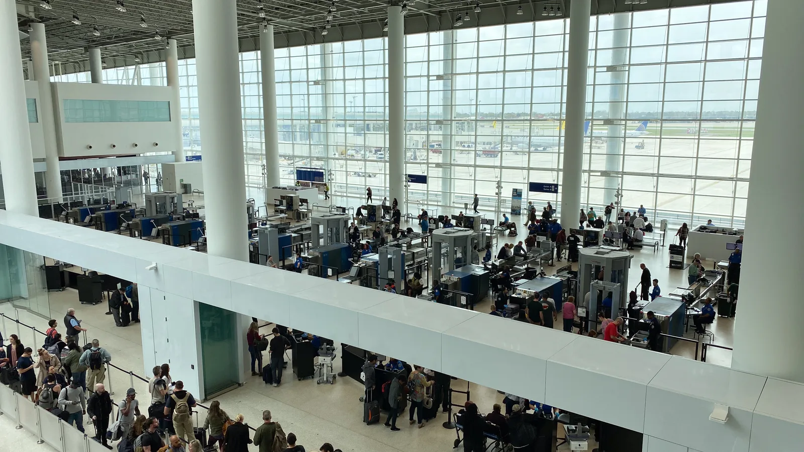 Travelers queue beside screening equipment at an airport security checkpoint.