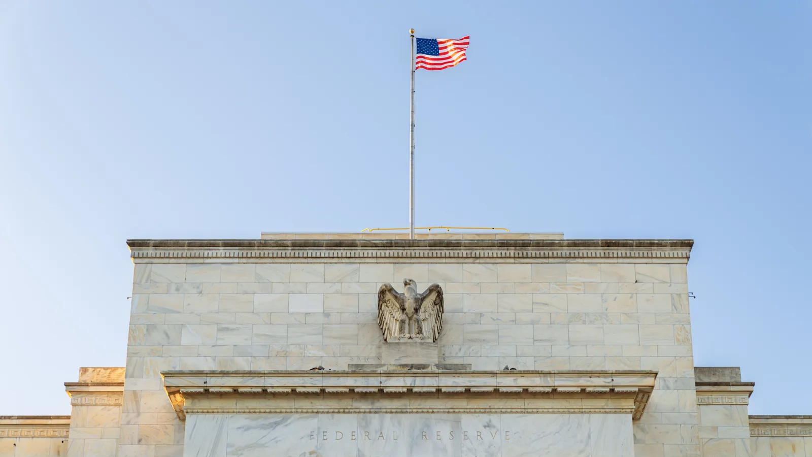 The pale stone Federal Reserve building stands behind a lawn and trees under a bright sky.