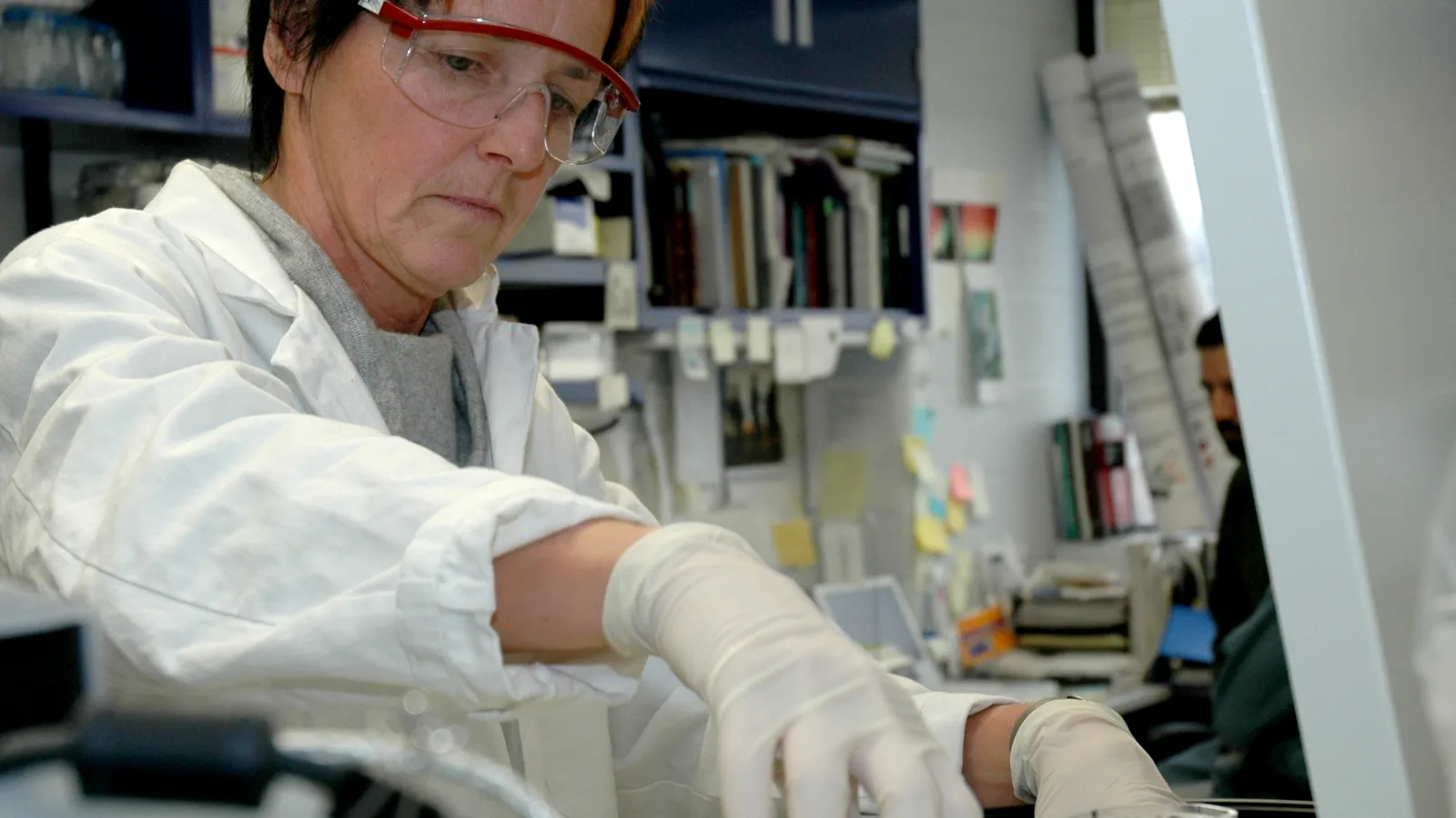 A laboratory scientist in protective clothing works with instruments on a crowded research bench.