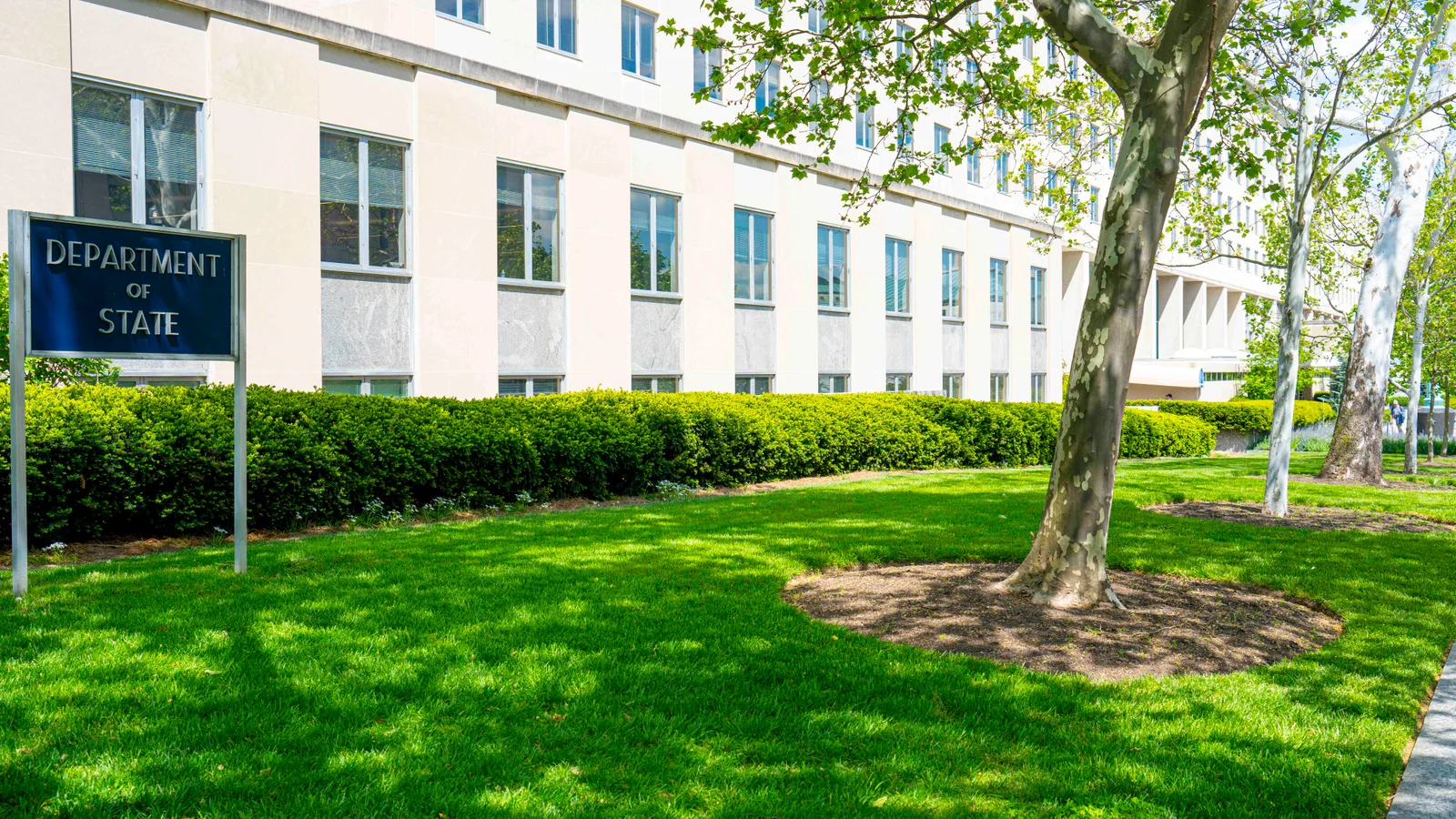 The State Department’s long stone headquarters fills the frame behind trees and a broad sidewalk in Washington.