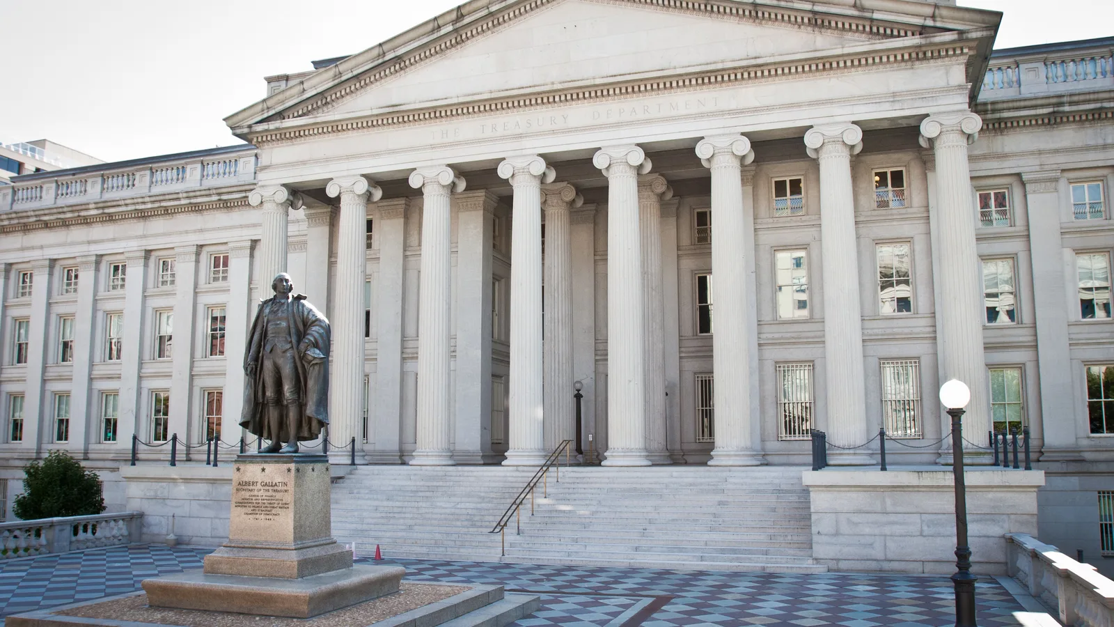 The colonnaded United States Treasury Building stands behind a statue and winter trees.