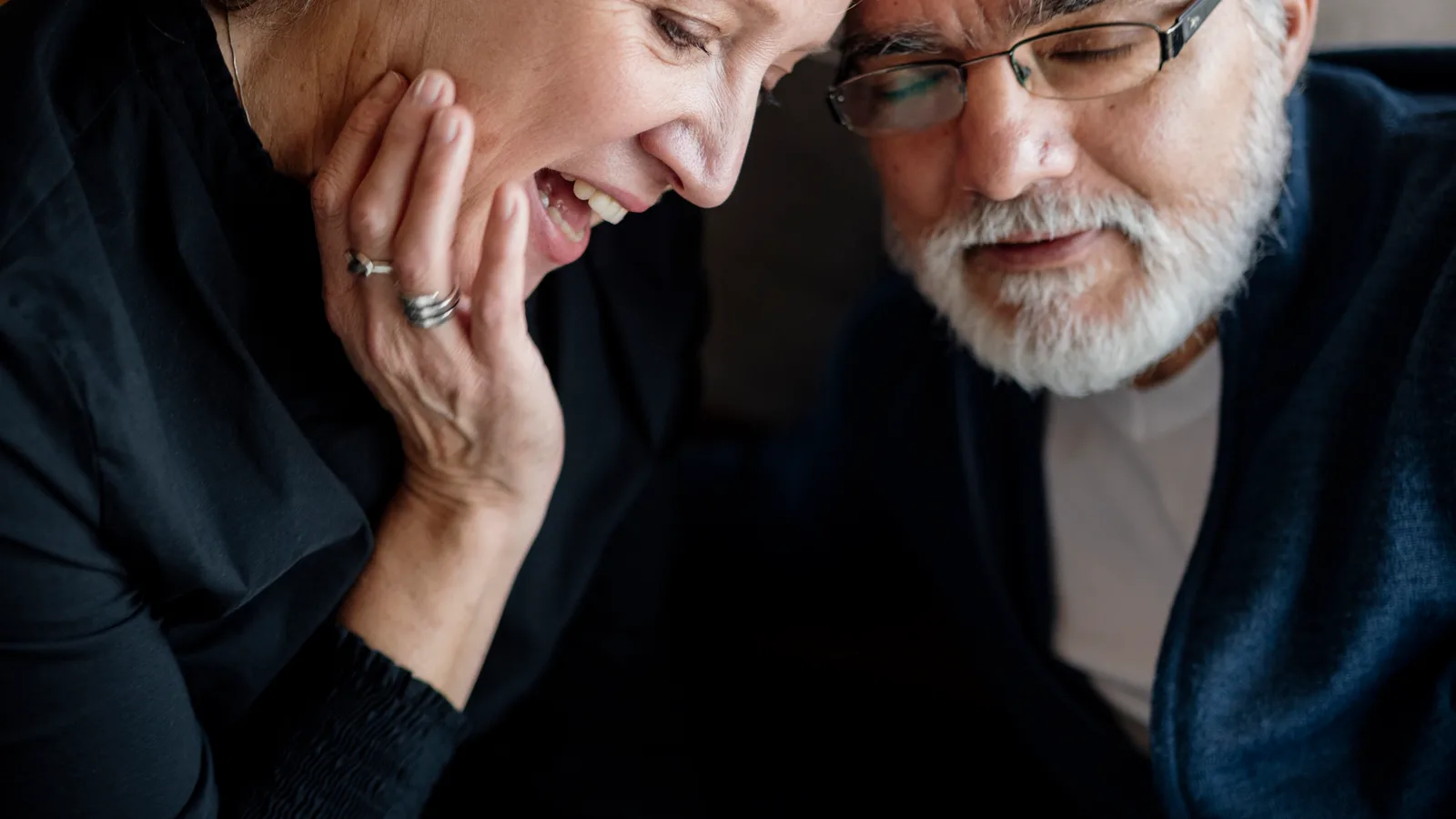 An older couple sits closely together reading a book at home.