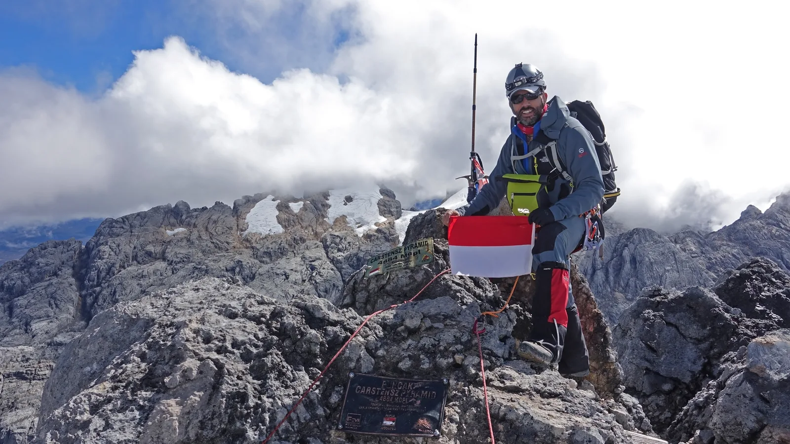 Climber at the rocky Puncak Jaya summit with clouds and patches of tropical ice behind him.
